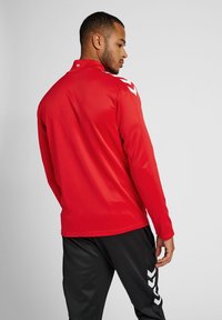 Rød sportsbluse med lange ærmer og høj krave samt hvide detaljer på skuldrene. Glat, letvægtsstof; parret med sorte underdele.