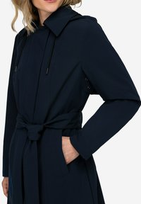 Námorně modrý trenčkot s opaskem, kapucí a bočními kapsami; má elegantní design s hladkou texturou a decentním stylem.