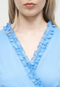 Jauna moteris dėvi šviesiai mėlyną palaidinę su raukinukais puošta V formos apykakle, matomas jos apatinis veidas, kaklas ir viršutinė krūtinės dalis.