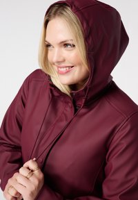 Veste imperméable bordeaux avec capuche, présentant une texture lisse, une fermeture éclair et des boutons-pression. Mains serrées autour de la fermeture éclair.