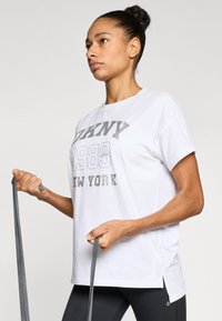 Άσπρο βαμβακερό t-shirt με κοντά μανίκια, διακοσμημένο με ασημένιο γραφικό τύπωμα "DKNY 1989 NEW YORK" και πλαϊνές σχισμές. Φοριέται με μαύρο κολάν.