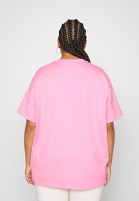 Person med flätat hår, bärande hängande örhängen, en lös rosa t-shirt och ljusa byxor, stående mot en enkel bakgrund.