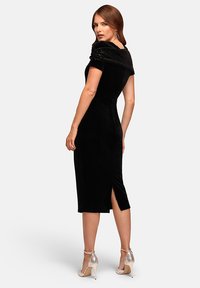 Donna che indossa un abito midi in velluto nero aderente con dettaglio off-shoulder in paillettes, spacco sul retro e scarpe con tacco alto e cinturino alla caviglia di colore argento.