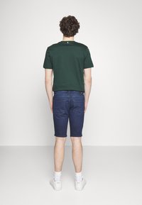 Osoba s kudrnatými vlasy se otáčí zády, má na sobě zelené tričko, modré kraťasy a bílé tenisky, na jednotném světlém pozadí.