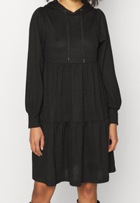 En person bär en svart, lager-på-lager klänning med en huva och långa ärmar. Klänningen har en avslappnad passform och en volangkjol.