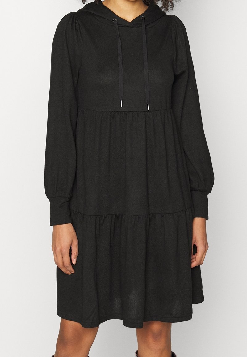 En person bär en svart, lager-på-lager klänning med en huva och långa ärmar. Klänningen har en avslappnad passform och en volangkjol.