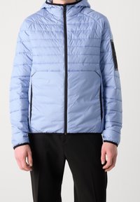 Ljusblå puffjacka med huva, med horisontella quiltade linjer, svart dragkedja och sidofickor; svarta accenter på sidorna.