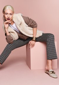 Faux-Fur-Weste in Creme über einem braunen Cardigan, kombiniert mit gemusterten Schwarz-Weiß-Hosen und beigen Loafers mit goldenen Akzenten.