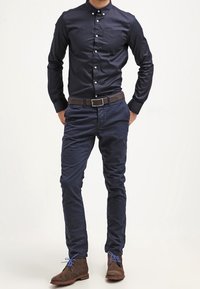 Námořnická košile na knoflíky, námořnické chinos kalhoty, hnědý opasek a hnědé boty s modrými šňůrkami. Rovný střih, hladký materiál, žádné viditelné vzory ani akcenty.