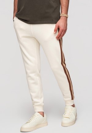 Homme portant un pantalon de jogging blanc avec deux bandes brunes sur le côté, des baskets blanches et une chemise vert olive foncé, debout les mains dans les poches.
