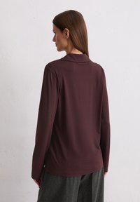 Ilgi piedurkņu, dziļi bordo tops ar mīksti strukturētu apkakli, izgatavots no gludas auduma, kopā ar pelēkiem platām biksēm.