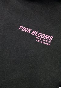 Zwarte stof met roze tekst, "PINK BLOOMS," met de tagline, "DE KUNST VAN VREDE & EEN ONGEDEZELD GEEST," in een vet, eenvoudig lettertype.