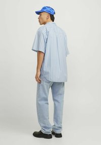 Joven vestido con una camisa corta de rayas azules, jeans azul claro, zapatos negros y una gorra de visera azul brillante, de pie contra un fondo plano.