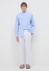 Camicia button-up a righe blu chiaro con colletto a band, abbinata a pantaloni bianchi in lino e infradito marroni. Tessuto texturizzato e vestibilità rilassata.