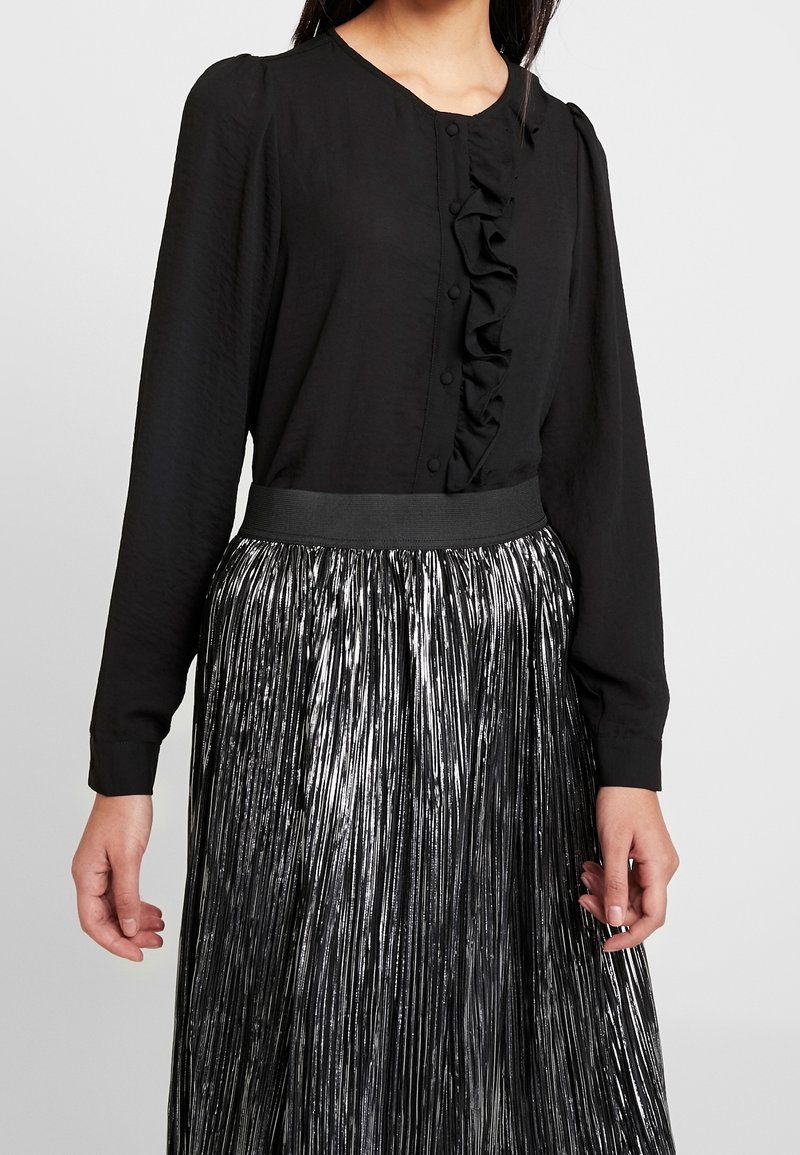 Femme portant un chemisier noir à manches longues avec des boutons à volants sur le devant et une jupe plissée argentée brillante avec une ceinture noire.