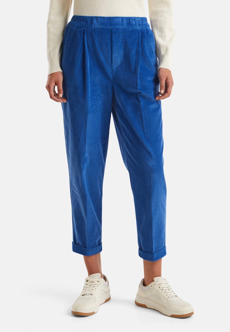 Pantalones de pana azul con un corte relajado, que presentan pliegues frontales, puños enrollados y una cinturilla elástica, combinados con zapatillas blancas.