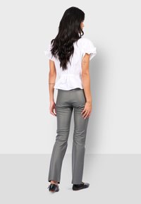 Vrouw met lang donker haar, gekleed in een wit overhemd met korte mouwen, grijze skinny pants, zwarte schoenen, staande met de rug naar de camera tegen een effen achtergrond.