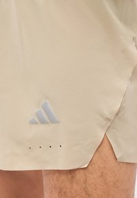 Μπεζ σορτς με γκρι λογότυπο Adidas και μικρές οπές αερισμού, που φοριούνται από άτομο που δείχνει μέρος του τριχωτού μηρού του.