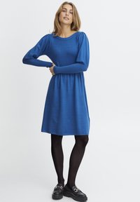 Blå stickad klänning med långa, puffärmar, rund halsringning och en åtsittande midja, som sträcker sig ner till knäna. Bärs med svarta strumpbyxor och loafers.