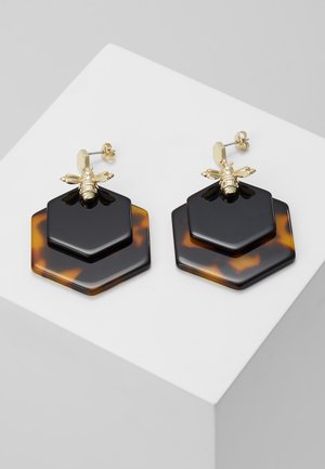 Złote kolczyki w kształcie pszczół przymocowane do warstwowych, czarnych i żółwicznych sześciokątnych zawieszek na białym tle.