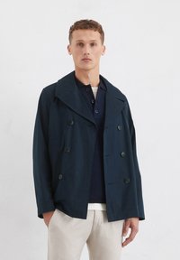 Marineblå dobbeltbrystet jakke, bred krave, forlommer og knapdetaljer. Båret over en struktureret mørk bluse og en off-white nederdel.