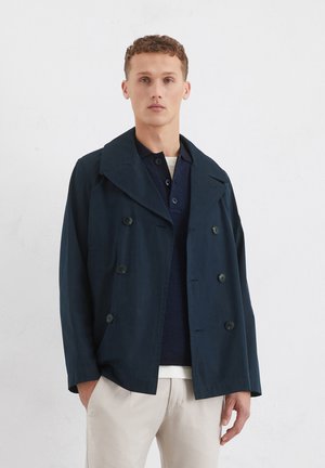 Marineblå dobbeltbrystet jakke, bred krave, forlommer og knapdetaljer. Båret over en struktureret mørk bluse og en off-white nederdel.