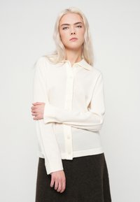 Mujer joven con cabello rubio largo, vistiendo un cárdigan blanco abotonado y una falda oscura, de pie contra un fondo claro y liso.