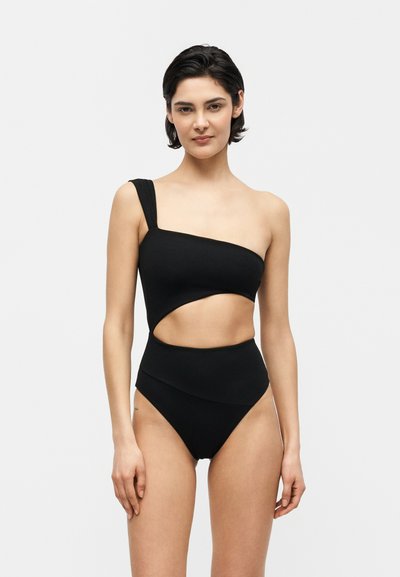 Femme portant un maillot de bain noir une épaule avec une découpe à la taille, debout devant un fond gris clair uni.