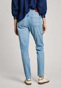 Šviesiai mėlyni džinsai su tiesiu įpjovimu, dviem užpakalinėmis kišenėmis, šiek tiek suplėšytais kraštais, derinami su baltais sportbačiais, papuoštais gyvūnų raštų akcentais.