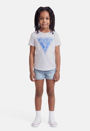 Mladé dievča s vrkočmi, oblečené v bielom tričku s modrým logom "GUESS", svetlomodrých rifľových šortkách, bielych ponožkách a bielych teniskách stojí.