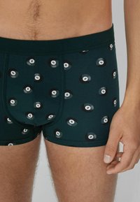 Männlicher Oberkörper in dunkelgrünen Boxerbriefs mit schwarz-weißen 8-Ball-Motiven, der vor einem neutralen Hintergrund steht.