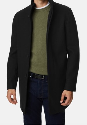 Homme portant un manteau noir boutonné par-dessus un pull vert olive et un jean bleu foncé, debout avec les mains détendues le long du corps.