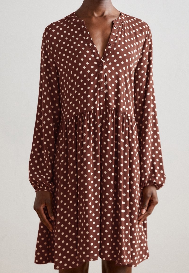 Vestido marrón con lunares de tela suave, con escote en V, mangas largas, botones y una cintura fruncida que ofrece una silueta fluida.