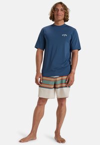 Rash guard de manga corta azul marino con logo; bañador de rayas multicolores en beige claro, verde, naranja y azul. Descalzo.