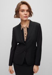 Black tailored blazer with a single button closure, notch lapels, and two front pockets, layered over a beige collared shirt.