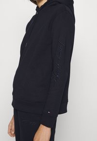 Sudadera con capucha azul marino con cordón, que presenta el logotipo bordado "Tommy Hilfiger" en azul oscuro a lo largo de la manga y puños acanalados. Textura suave.