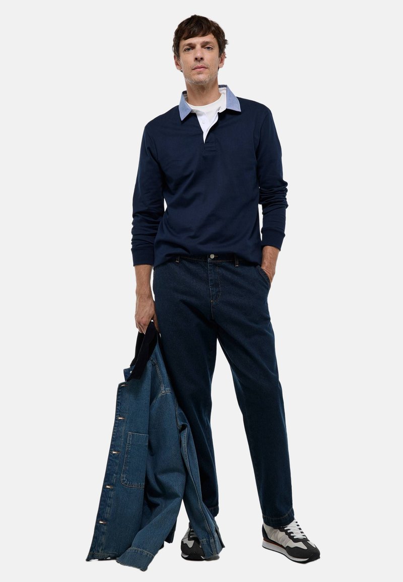 Polo à manches longues marine avec un col bleu clair, associé à un jean bleu foncé. Une veste en denim est accrochée à son bras gauche. Il porte des baskets grises et noires.