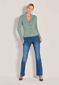 Eine Frau trägt eine hellgrüne, geknöpfte Strickjacke, blaue Jeans und dunkle Absätze und steht vor einem einfarbigen, hellen Hintergrund.