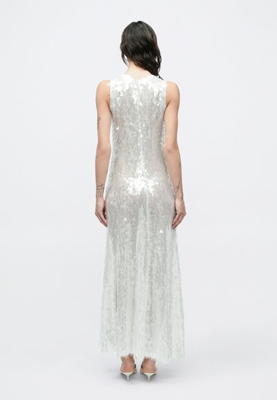 Sötét hajú nő, aki ujjatlan, földig érő, fehér flitteres ruhát és világos színű, alacsony sarkú cipőt visel, háttal áll egy egyszínű háttér előtt.