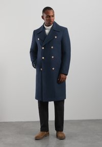 Marineblå dobbeltradet frakke i uldblanding med store guldknapper, sidelommer, brede revers, kombineret med en cremefarvet sweater og mørke bukser.