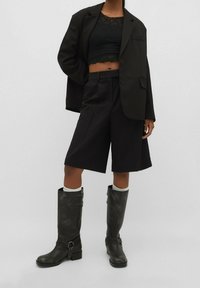 Zwarte oversized blazer, cropped top van kant, op maat gemaakte zwarte Bermuda-shorts en kniehoge zwarte laarzen met gespen en textuurelementen.