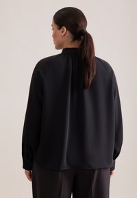 Blusa nera a maniche lunghe con vestibilità rilassata, caratterizzata da un colletto arricciato e una texture liscia. Dettaglio dei bottoni visibile ai polsini.