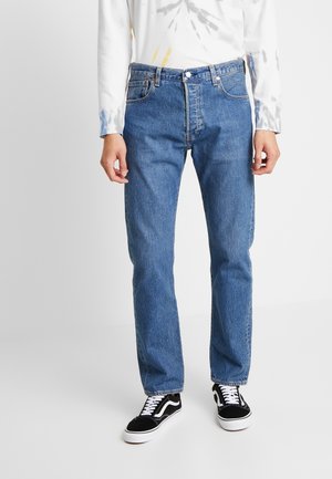 Person trägt blaue Jeans mit geradem Bein, schwarz-weiße Sneakers und ein weißes langärmliges Shirt mit dezenten Farbspritzern, steht auf einem grauen Boden.