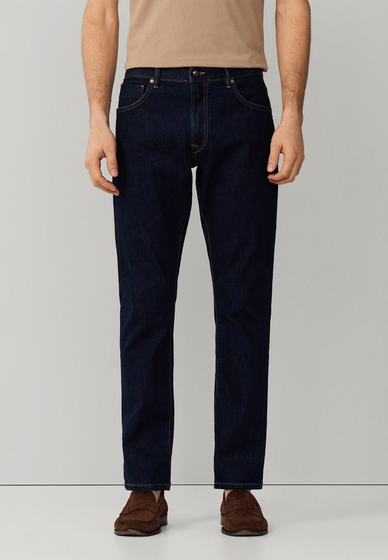 Muž oblečený v tmavomodrých džínsach s rovnými nohavicami, béžovej košeli a hnedých semišových mokasínach, stojaci pred jednoduchým svetlosivým pozadím.