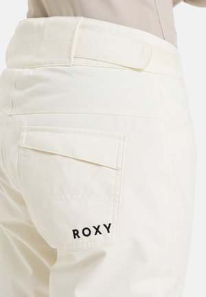 Cremefarvede bukser med en glat tekstur, der har en baglomme og et sort "ROXY" logo broderet på lommen.