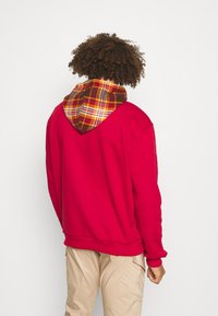 Sudadera roja con capucha forrada de cuadros en marrón. Presenta un corte holgado, puños y dobladillo acanalados. Costuras visibles en la parte posterior.