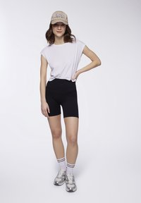 Shorts neri, t-shirt bianca a maniche corte, cappellino beige con testo "NEW YORK", sneakers argentate e calzini bianchi con strisce nere.