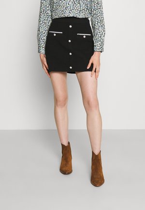 Mannequin portant une mini-jupe noire boutonnée devant avec des poches bordées de blanc, un chemisier à fleurs manches longues, et des bottines en daim marron.
