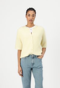 Gul kortermet cardigan laget av myk, strukturert stoff, med rund hals og knapplukking, parret med lyseblå jeans.