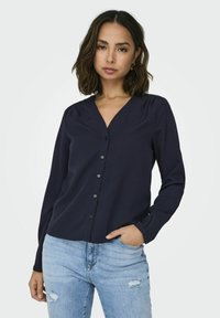 Chemise boutonnée bleu marine avec un col en V, des manches longues et une texture subtile. Associée à un jean déchiré bleu clair.